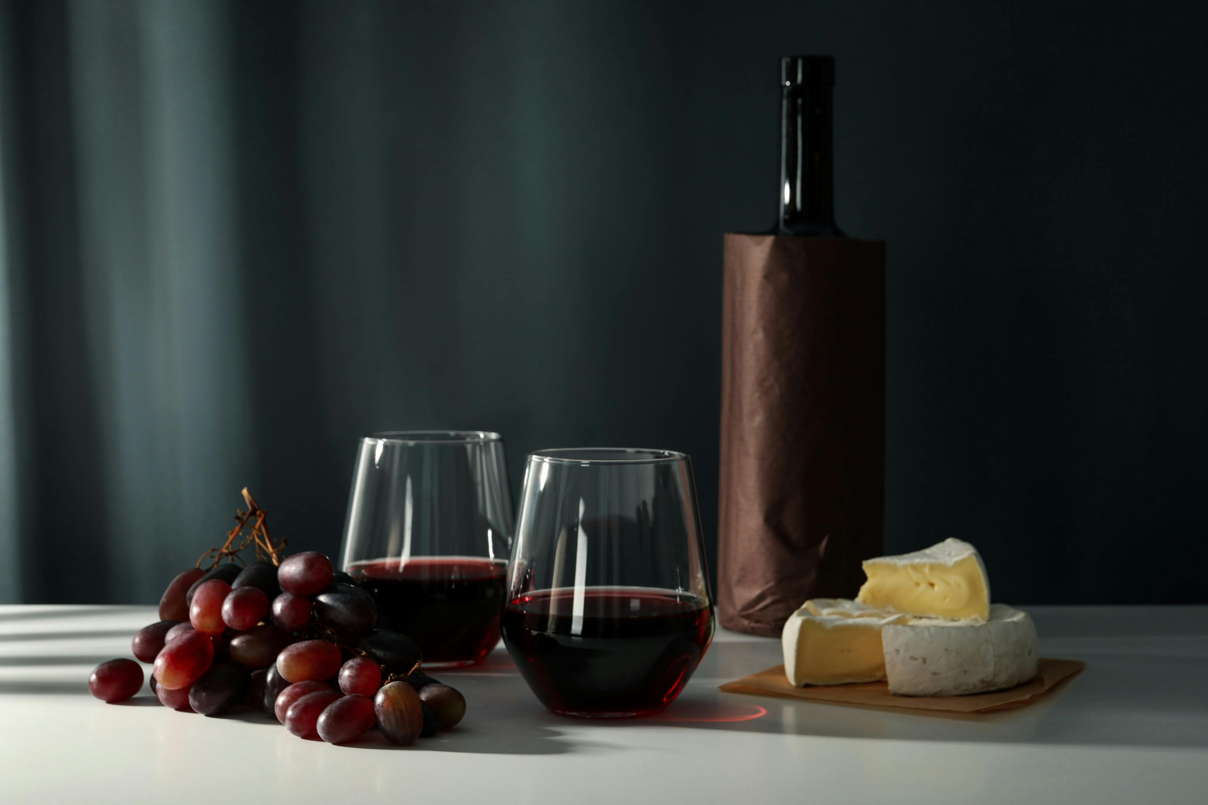 Vinflaska, glas med rött vin, druvor och en bit ost på ett bord mot en mörk bakgrund.